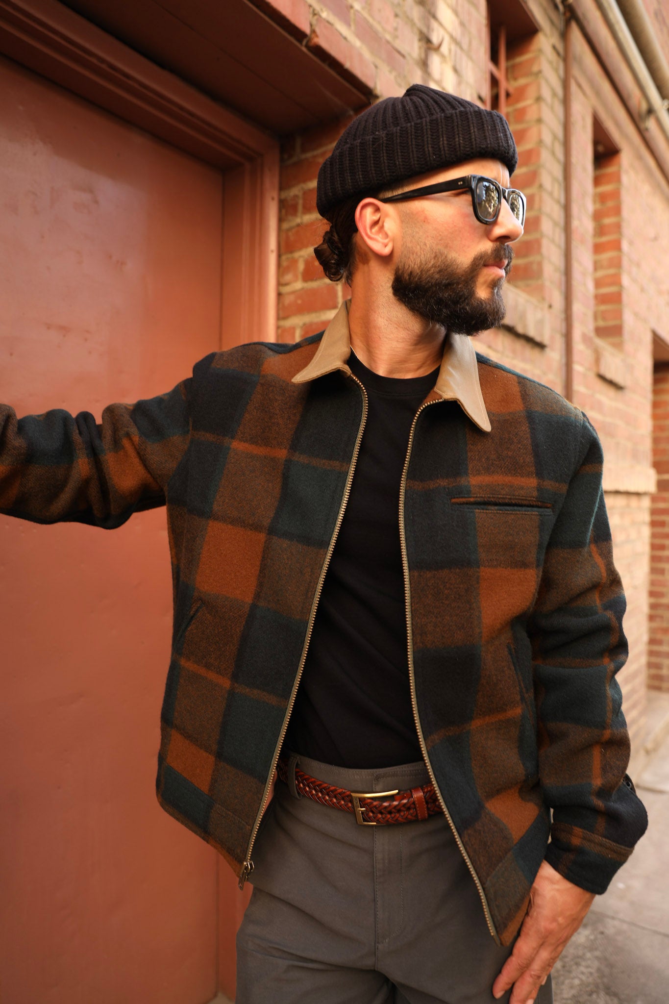 A man wearing a brown and dark green plaid overshirt jacket with a tan collar, styled with a black t-shirt, gray trousers, a black beanie, and sunglasses, standing outdoors against a brick wall and reddish-brown door.