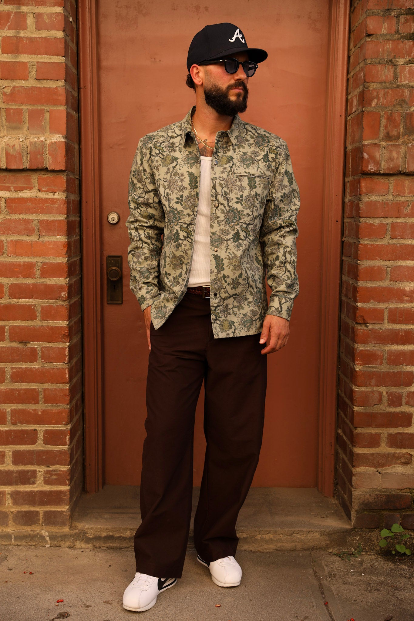 A man stands in front of a brick doorway wearing a green and beige floral patterned button-up shirt, dark brown pants, a black baseball cap with a white 'A' logo, sunglasses, and white sneakers.