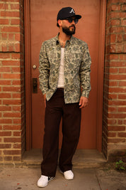 A man stands in front of a brick doorway wearing a green and beige floral patterned button-up shirt, dark brown pants, a black baseball cap with a white 'A' logo, sunglasses, and white sneakers.
