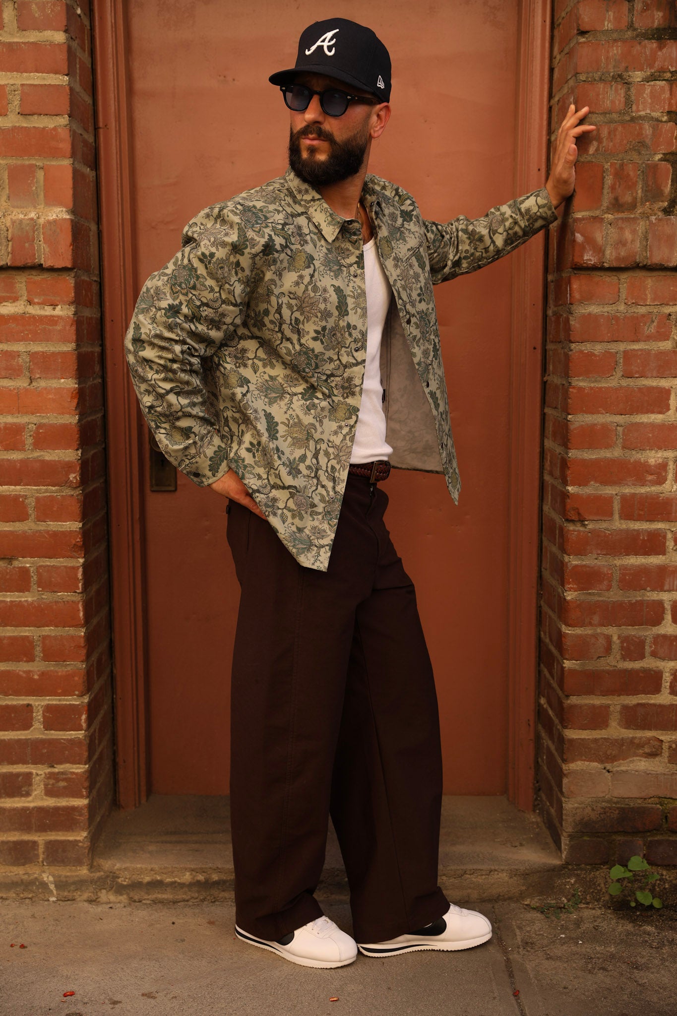 A man wearing a camouflage bomber jacket, dark brown wide-leg pants, a white t-shirt, and a black baseball cap with a white 'A' logo, posing in front of a brick doorway.
