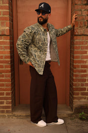 A man wearing a camouflage bomber jacket, dark brown wide-leg pants, a white t-shirt, and a black baseball cap with a white 'A' logo, posing in front of a brick doorway.