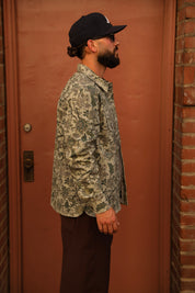 A man in profile wearing a black baseball cap, sunglasses, a long-sleeve button-up shirt with a green and beige floral camouflage pattern, and dark brown pants, standing in front of a reddish-brown wooden door.