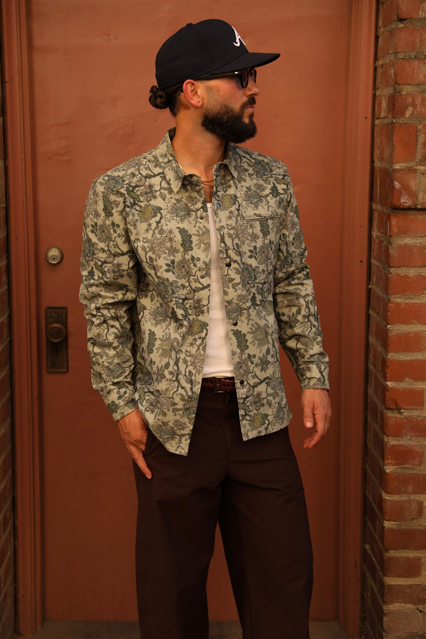 A man wearing a long-sleeve button-up shirt with a green and beige floral camouflage pattern, a white t-shirt, dark brown trousers, and a black baseball cap, standing in front of a reddish-brown wooden door.