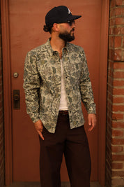 A man wearing a long-sleeve button-up shirt with a green and beige floral camouflage pattern, a white t-shirt, dark brown trousers, and a black baseball cap, standing in front of a reddish-brown wooden door.