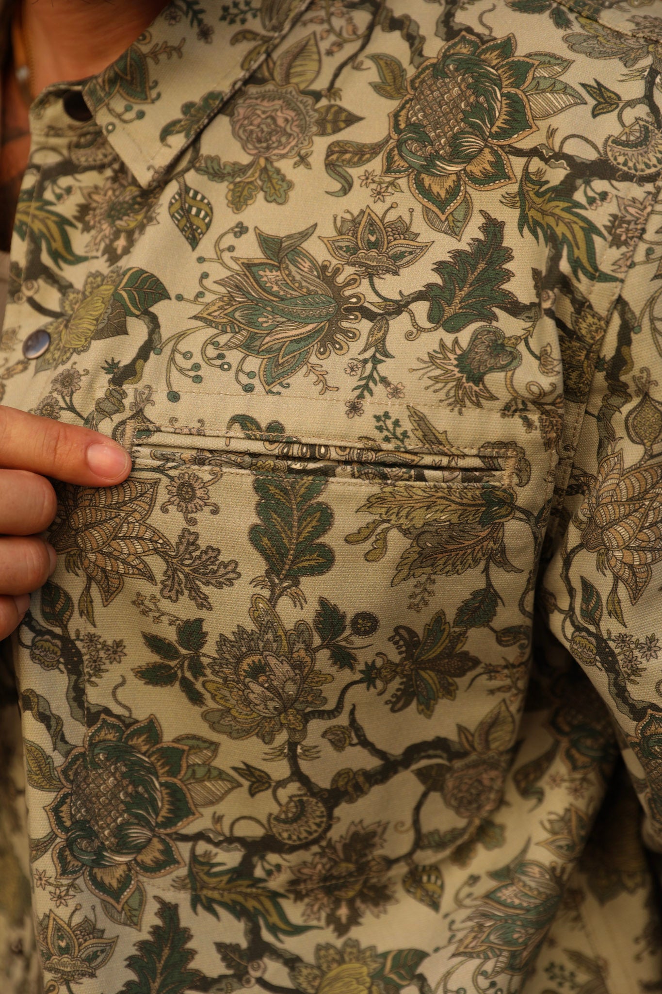 Close-up of a person wearing a beige button-up shirt with an intricate green and brown floral vine pattern, showing a hand holding the collar and a chest pocket.