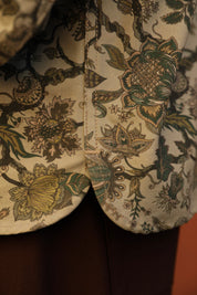 Close-up of a floral-patterned fabric garment with a cream base and green, olive, and mustard botanical print, shown with a dark brown lower section underneath.