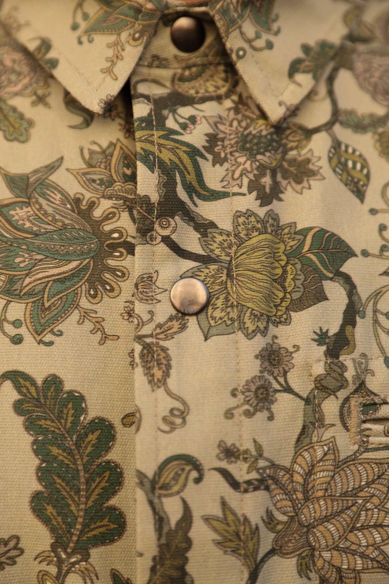Close-up of a button-up shirt collar and front placket featuring an intricate botanical and floral pattern in muted green, olive, tan, and brown tones on a light beige base fabric, with two metallic buttons visible.
