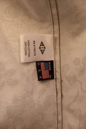Close-up of a garment's interior label showing '100% COTTON DRY-GEL' on a white tag and a black 'MADE IN USA' patch with an American flag, attached to light beige textured fabric.