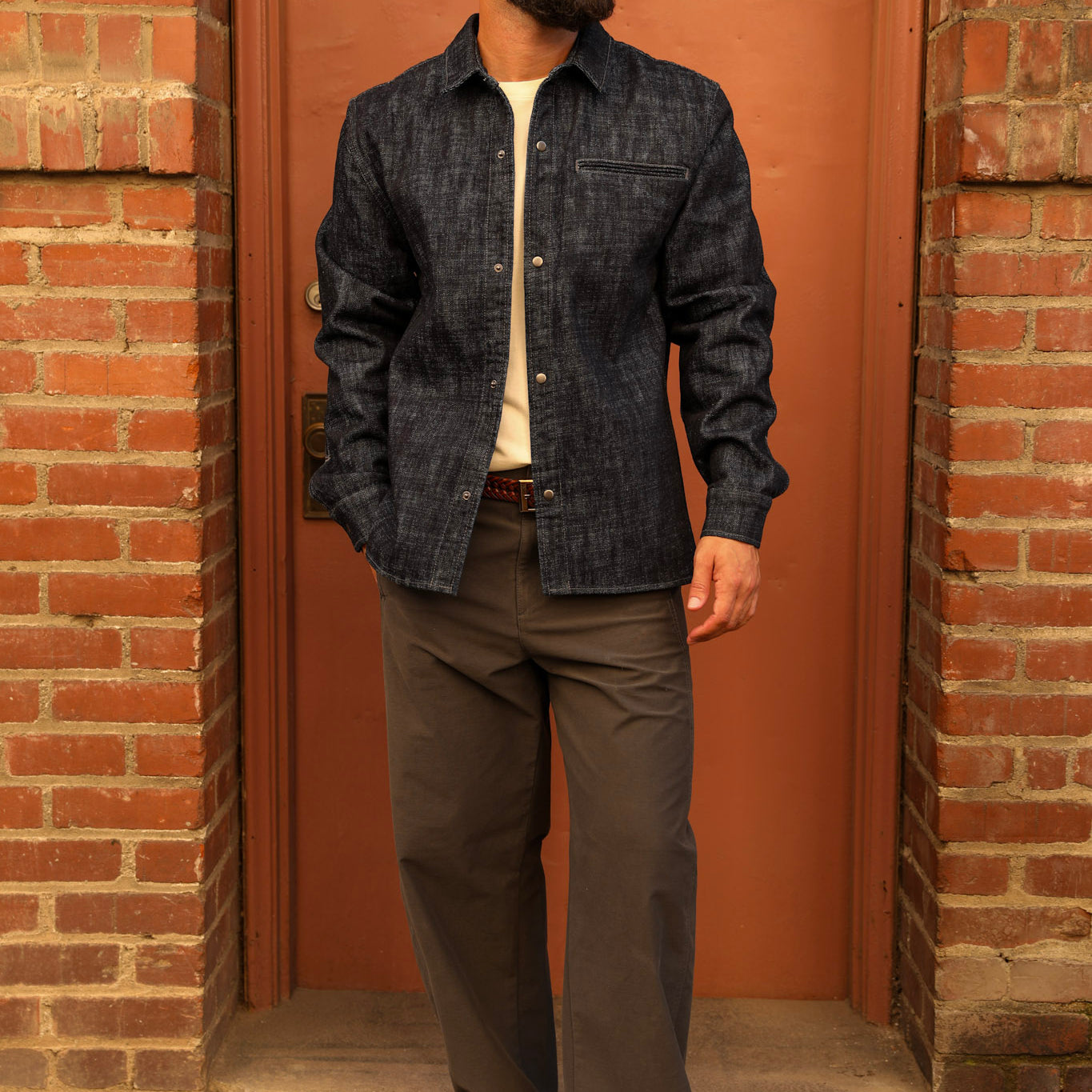 A man wearing a dark indigo denim button-down shirt, dark gray pants, white sneakers, and a black baseball cap with a white 'A' logo, standing in front of a rusted metal door framed by brick walls.