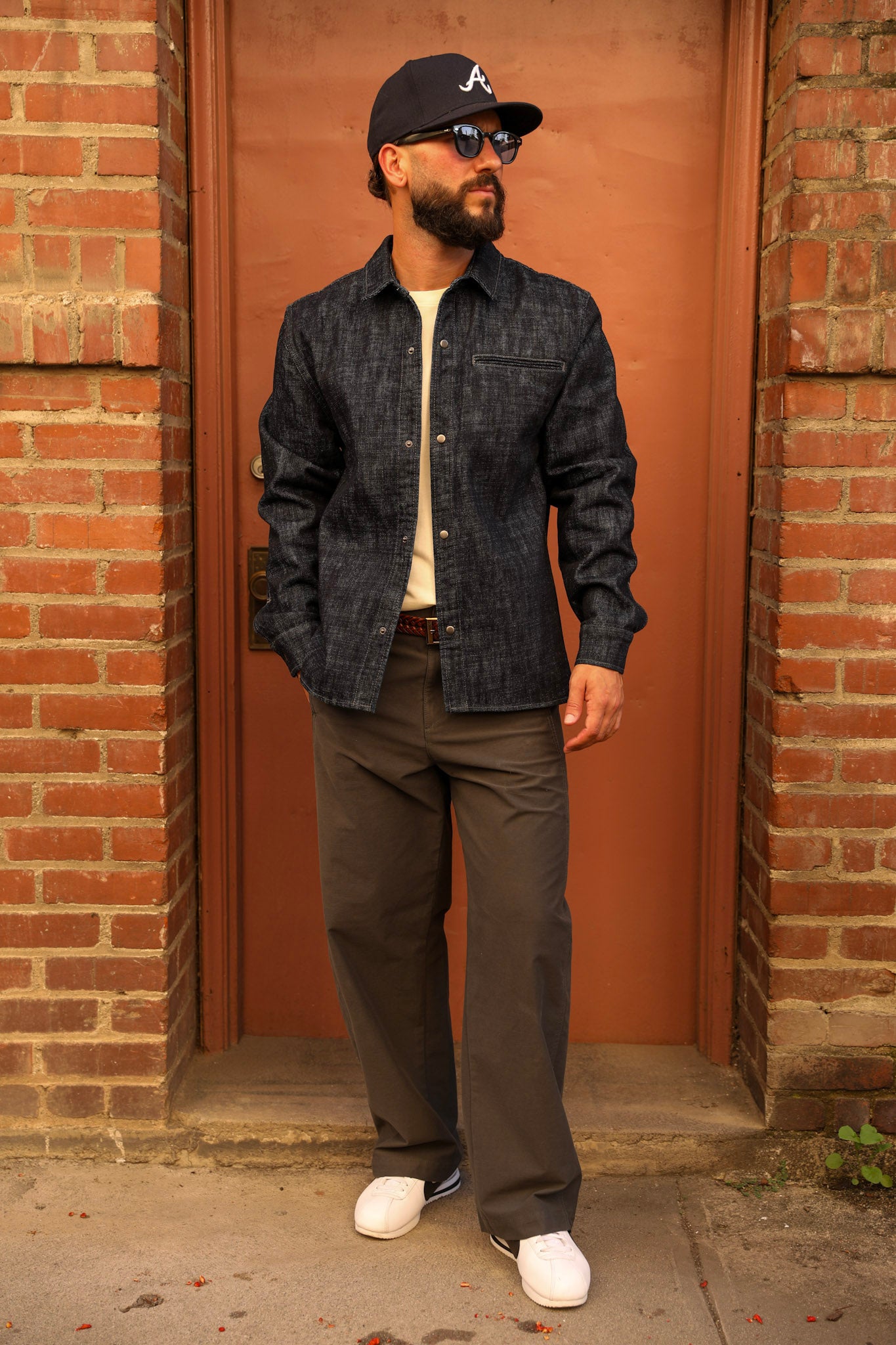 A man wearing a dark indigo denim button-down shirt, dark gray pants, white sneakers, and a black baseball cap with a white 'A' logo, standing in front of a rusted metal door framed by brick walls.