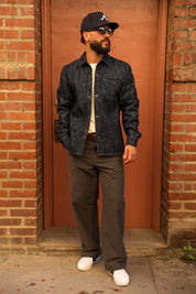 A man wearing a dark indigo denim button-down shirt, dark gray pants, white sneakers, and a black baseball cap with a white 'A' logo, standing in front of a rusted metal door framed by brick walls.
