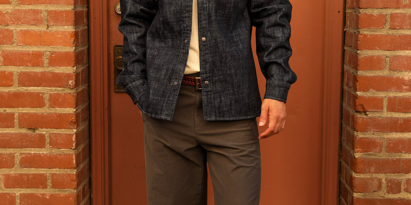 A man wearing a dark indigo denim button-down shirt, dark gray pants, white sneakers, and a black baseball cap with a white 'A' logo, standing in front of a rusted metal door framed by brick walls.