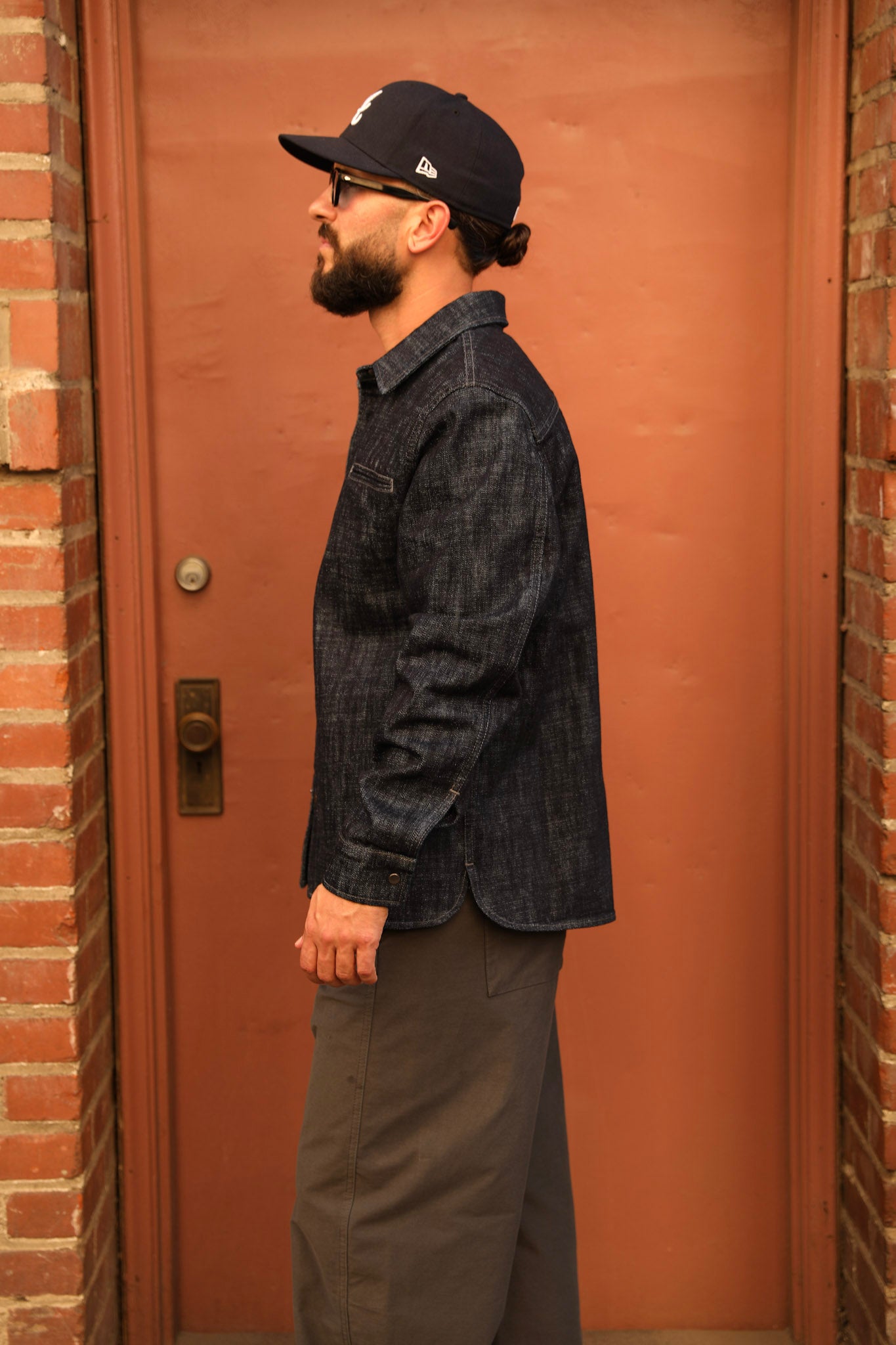 A man wearing a dark indigo denim shirt, dark gray pants, a black baseball cap, and glasses stands in profile against a warm terracotta-colored door framed by brick walls.