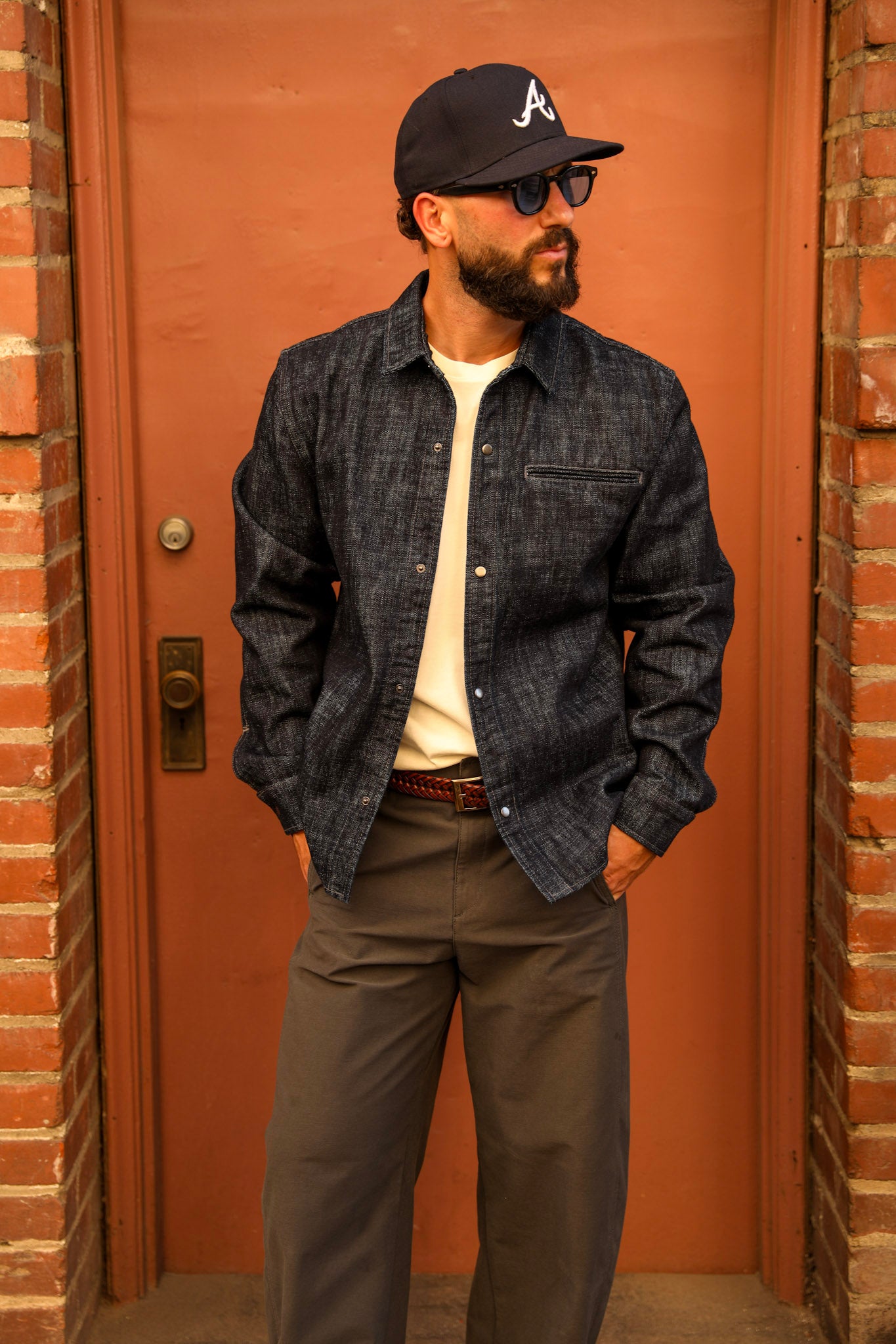 A man wearing a dark indigo denim button-up overshirt, olive pants, a cream t-shirt, sunglasses, and a black baseball cap with a white 'A' logo, standing in front of a rustic orange-brown door framed by brick walls.