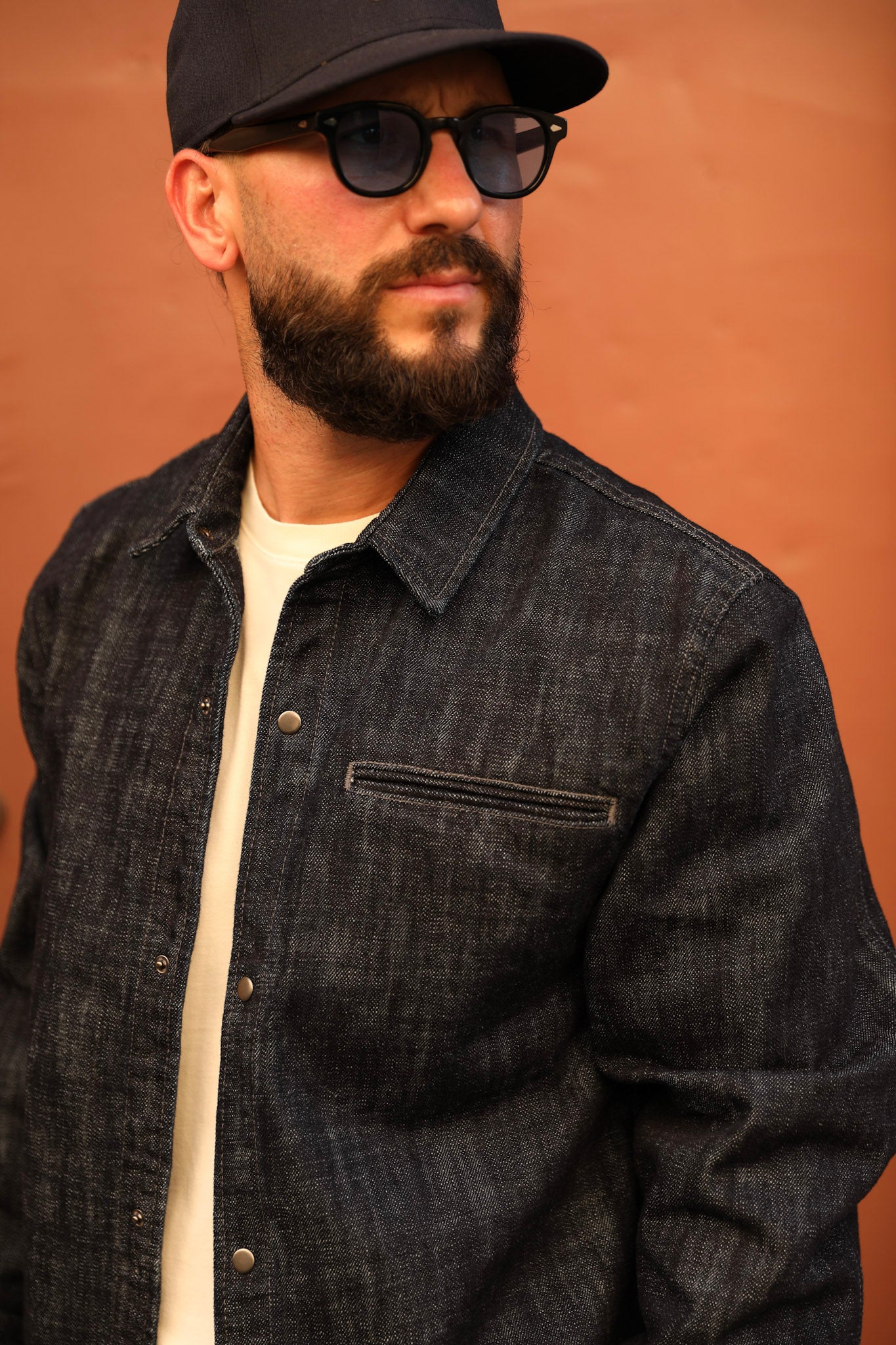 A man wearing a dark indigo denim textured overshirt over a cream t-shirt, a black baseball cap, and black sunglasses, set against a solid terracotta-colored background.