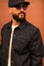 A man wearing a dark indigo denim textured overshirt over a cream t-shirt, a black baseball cap, and black sunglasses, set against a solid terracotta-colored background.