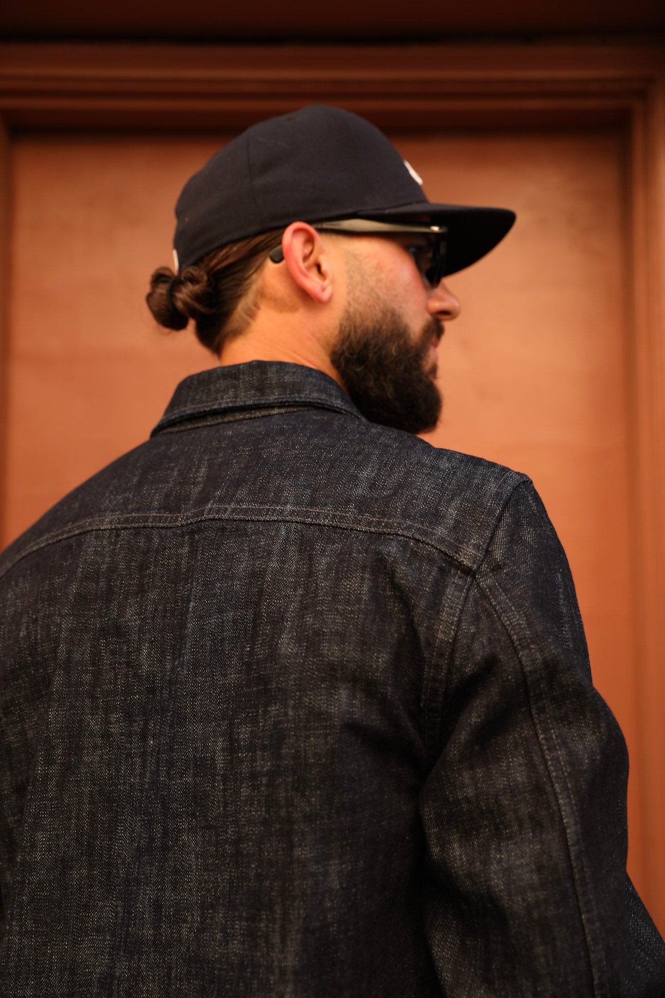A man shown from behind wearing a dark indigo denim jacket, a black baseball cap, and sunglasses, standing in front of a warm brown wooden door.