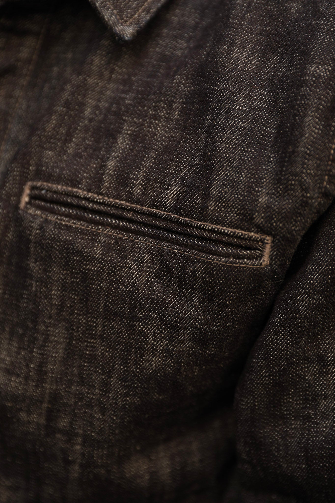 Close-up detail of a dark indigo textured fabric denim jacket, showing a horizontal welt pocket with double-stitched trim.