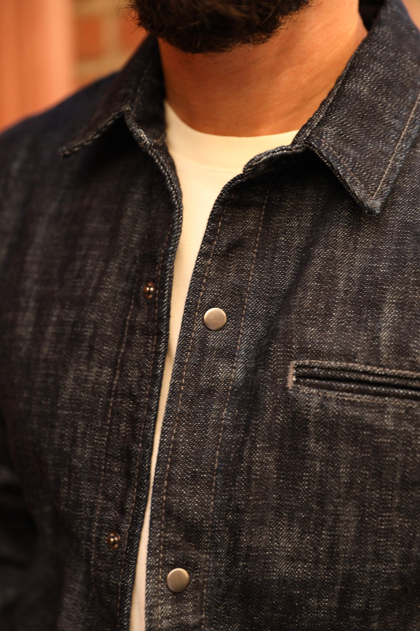 Close-up of a person wearing a dark indigo denim overshirt over a white t-shirt, showing the collar, front buttons, and chest pocket.