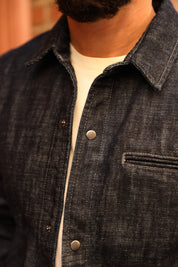 Close-up of a person wearing a dark indigo denim overshirt over a white t-shirt, showing the collar, front buttons, and chest pocket.
