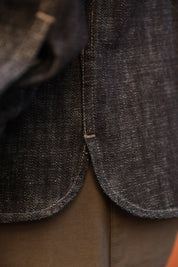 Close-up detail of a dark indigo denim garment, showing the raw hem, contrast stitching, and a portion of olive brown pants underneath.