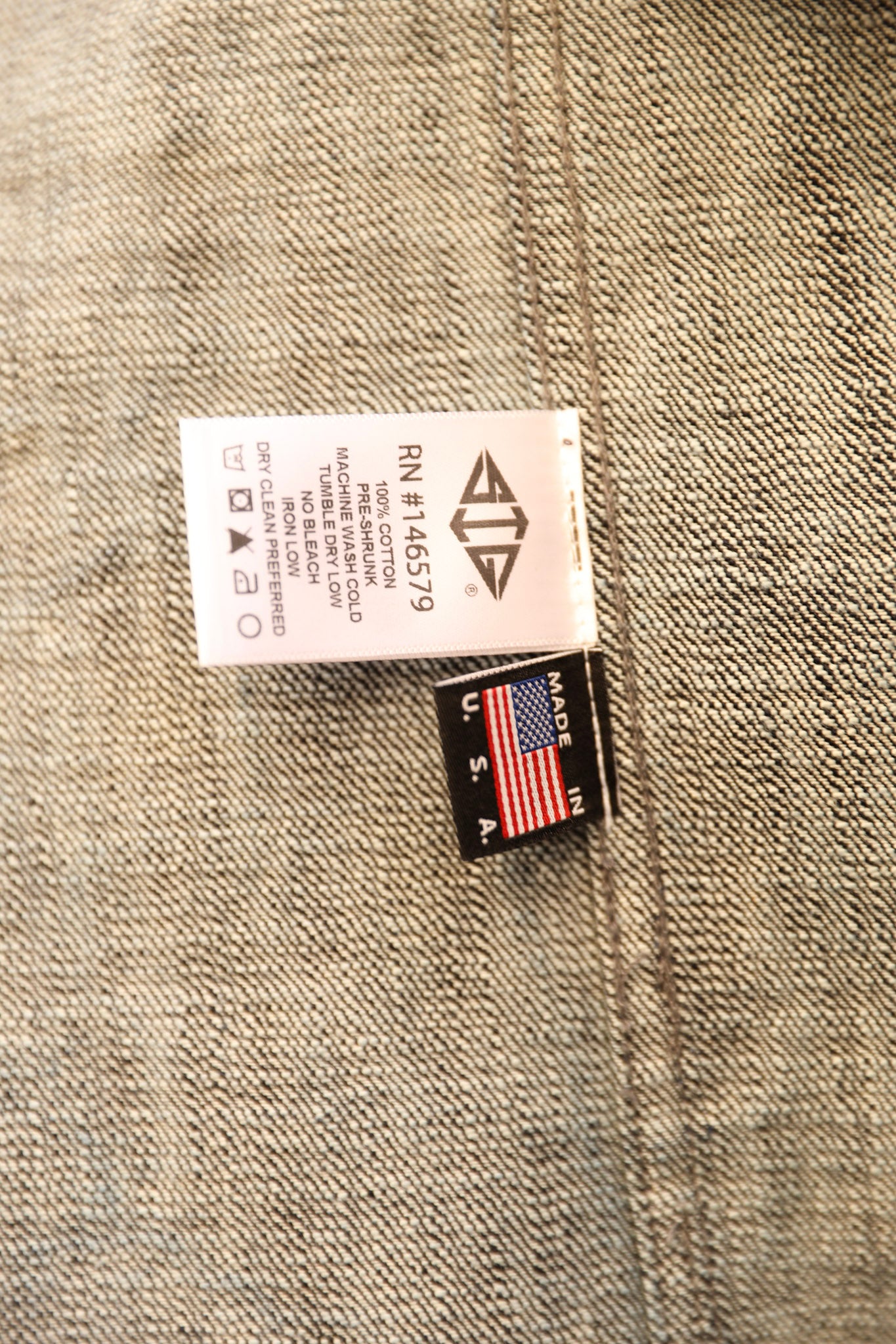 Close-up of a garment tag sewn onto beige woven fabric. The white rectangular size tag shows text including 'RN-414579' and a diamond-like logo, and the smaller black tag below it reads 'MADE IN USA' with an American flag.