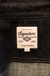 Close-up of a denim garment's inner label area showing a white woven tag with the brand name 'Signature' and 'MADE IN THE USA' above it, with a small size 'M' tab below.