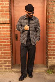 A man wearing a black knit beanie, black sunglasses, a black and gray plaid button-down shirt over a black t-shirt, black trousers, and black leather shoes, standing in front of a reddish-brown door framed by brick walls.
