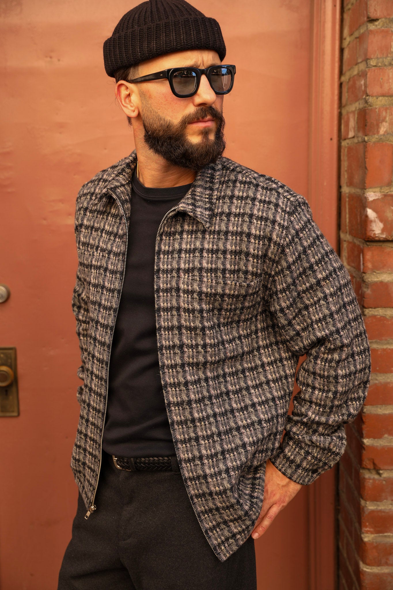 A man wearing a black knit beanie, black sunglasses, a black t-shirt, and a gray and black plaid overshirt jacket, standing in front of a terracotta-colored door and brick wall.