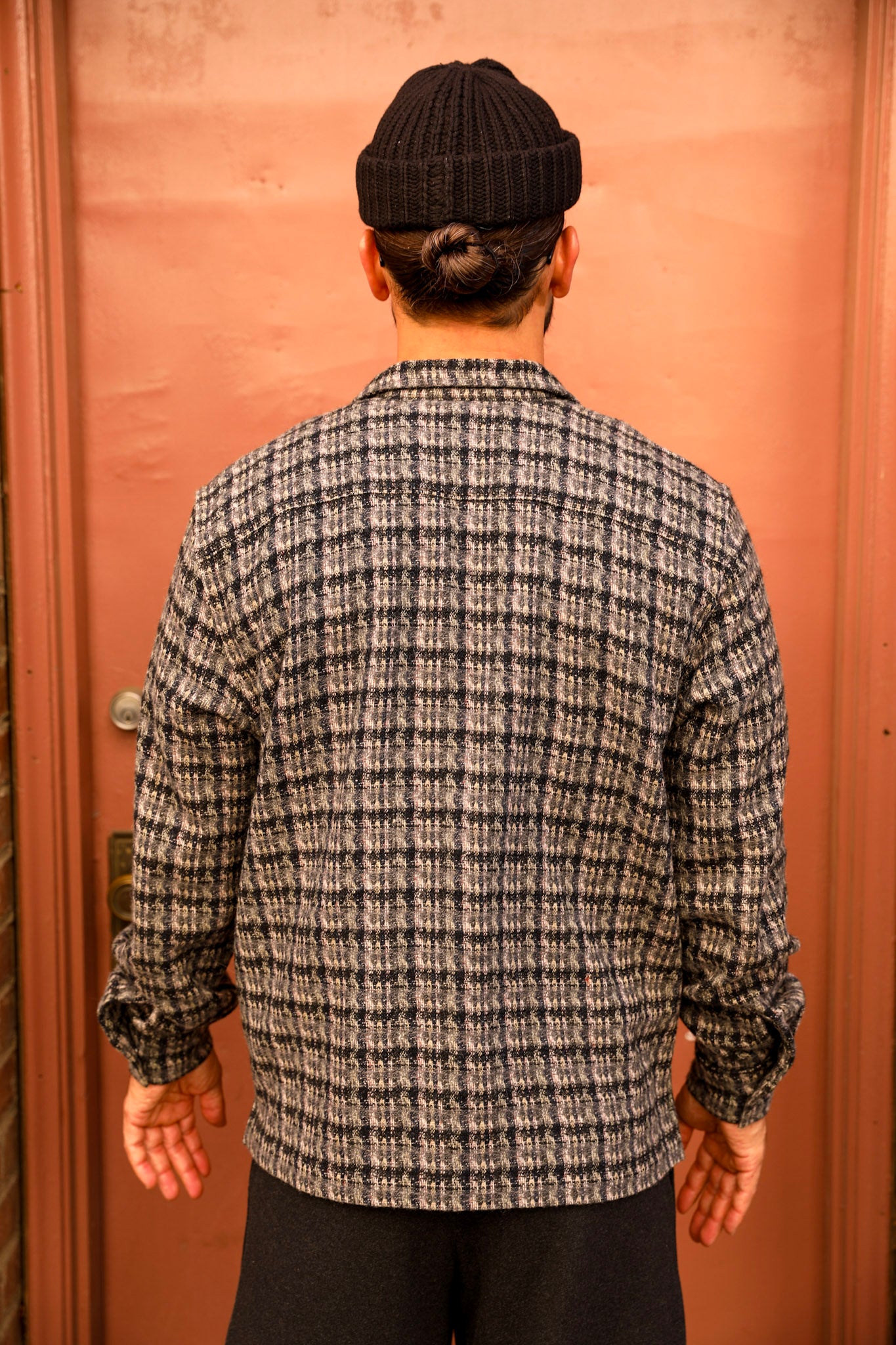 A man viewed from behind wearing a black knit beanie, a gray and black plaid long-sleeve button-up shirt, and black pants, standing in front of a warm orange-brown door.