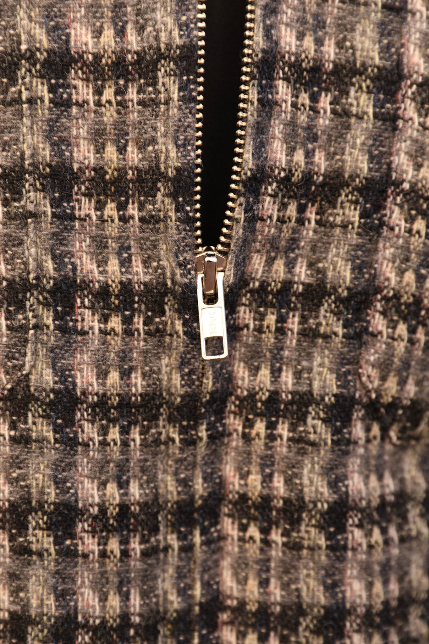 Close-up of a textured tweed zipper garment, showing a black, gray, and beige patterned fabric with a silver metal zipper pull.