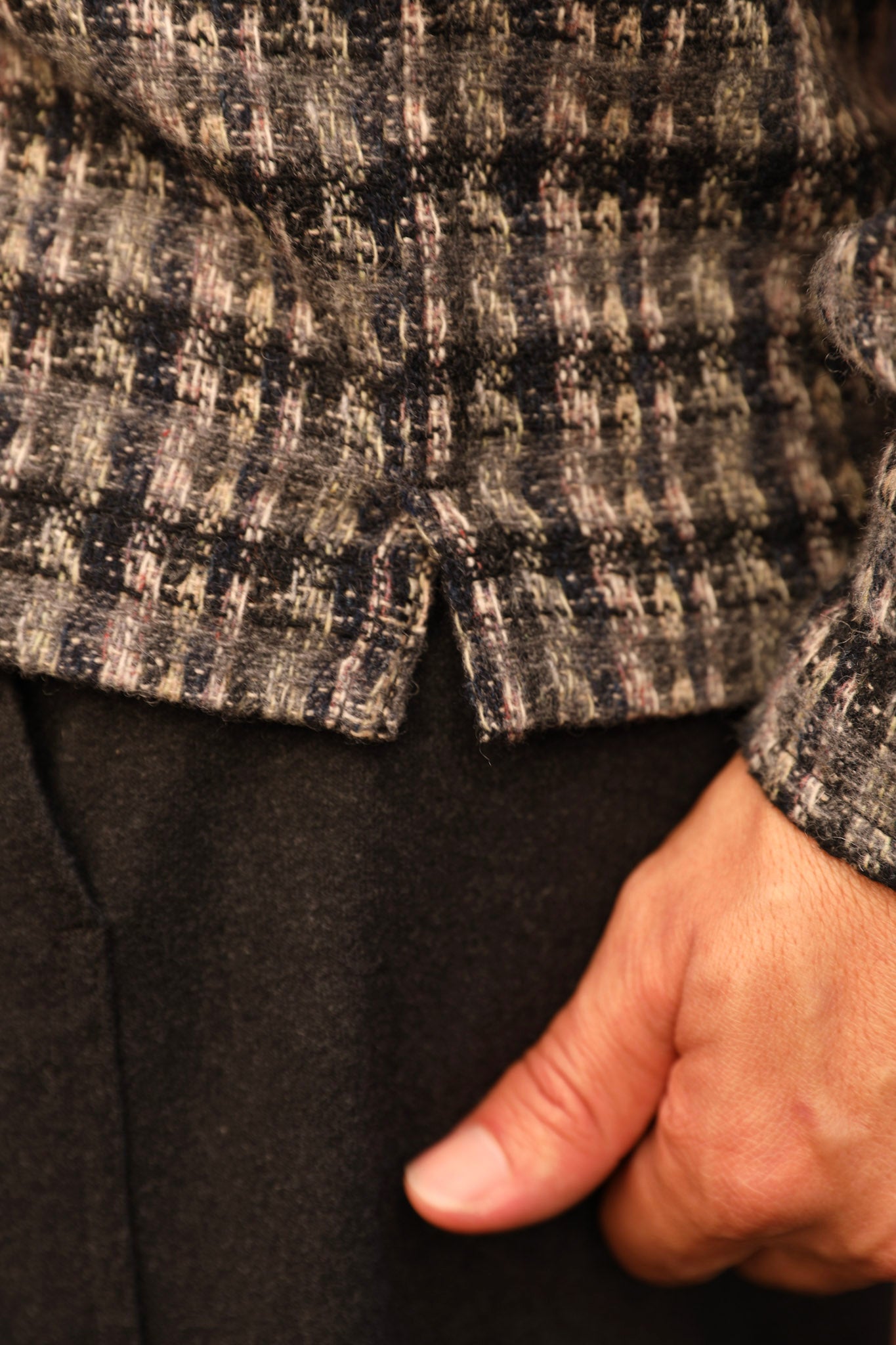Close-up of a person wearing a textured tweed jacket in black, beige, and gray tones, with a hand tucked into the pocket of dark trousers.