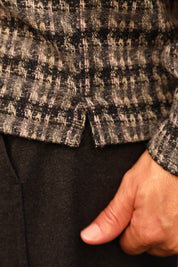 Close-up of a person wearing a textured tweed jacket in black, beige, and gray tones, with a hand tucked into the pocket of dark trousers.