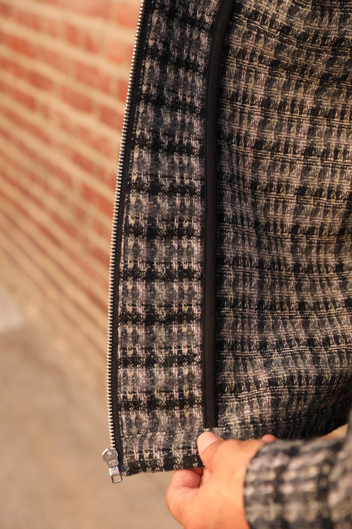 Close-up of a person's hand holding open a black zipper on a textured black and beige plaid tweed jacket, with a blurred brick wall in the background.