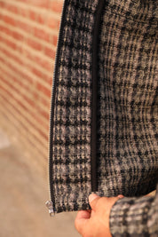 Close-up of a person's hand holding open a black zipper on a textured black and beige plaid tweed jacket, with a blurred brick wall in the background.