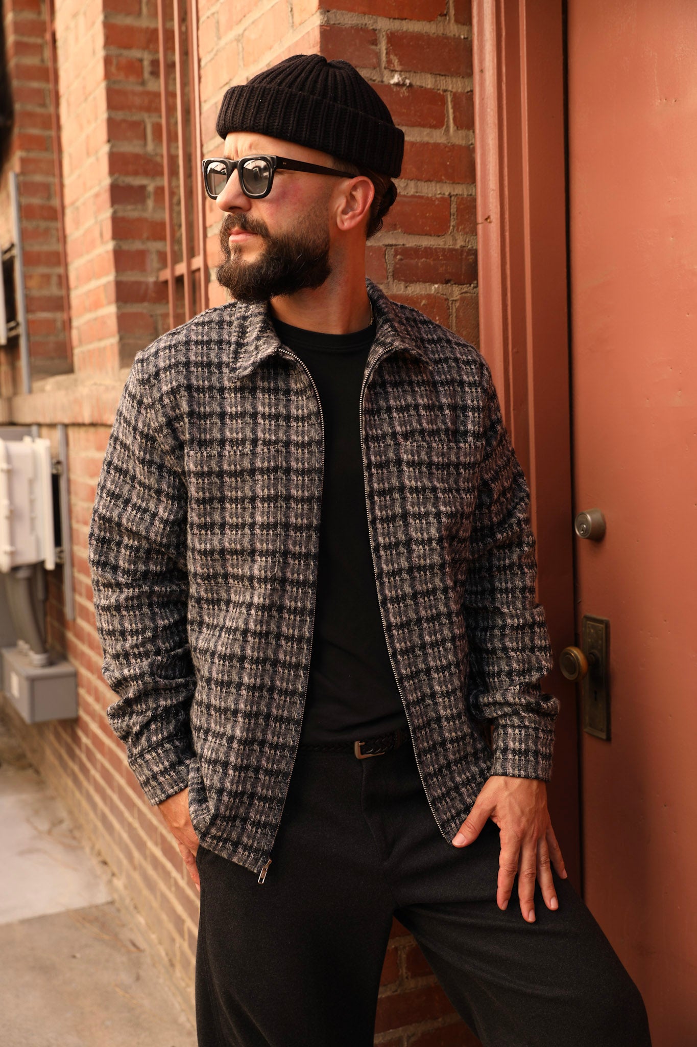 A man wearing a black knit beanie, black sunglasses, a black t-shirt, and a gray and black plaid zip-up overshirt, leaning against a brick wall next to a reddish-brown door.