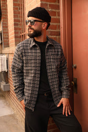 A man wearing a black knit beanie, black sunglasses, a black t-shirt, and a gray and black plaid zip-up overshirt, leaning against a brick wall next to a reddish-brown door.