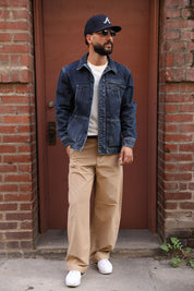 A man wearing a dark blue denim jacket, beige pants, white sneakers, a white t-shirt, and a navy baseball cap with a white 'A' logo, standing in front of a brown door framed by brick walls.