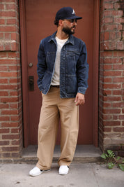 A man wearing a dark blue denim jacket, beige pants, white sneakers, and a navy baseball cap stands in front of a brown door framed by brick walls.