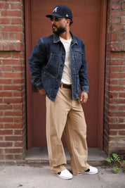 A man wearing a dark blue denim jacket, beige wide-leg trousers, a white t-shirt, white sneakers, and a navy baseball cap with a white 'A' logo, standing in front of a brown door framed by brick walls.