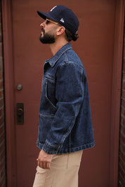 A man in profile wearing a dark blue denim jacket, beige pants, a navy baseball cap, and sunglasses, standing in front of a reddish-brown wooden door.