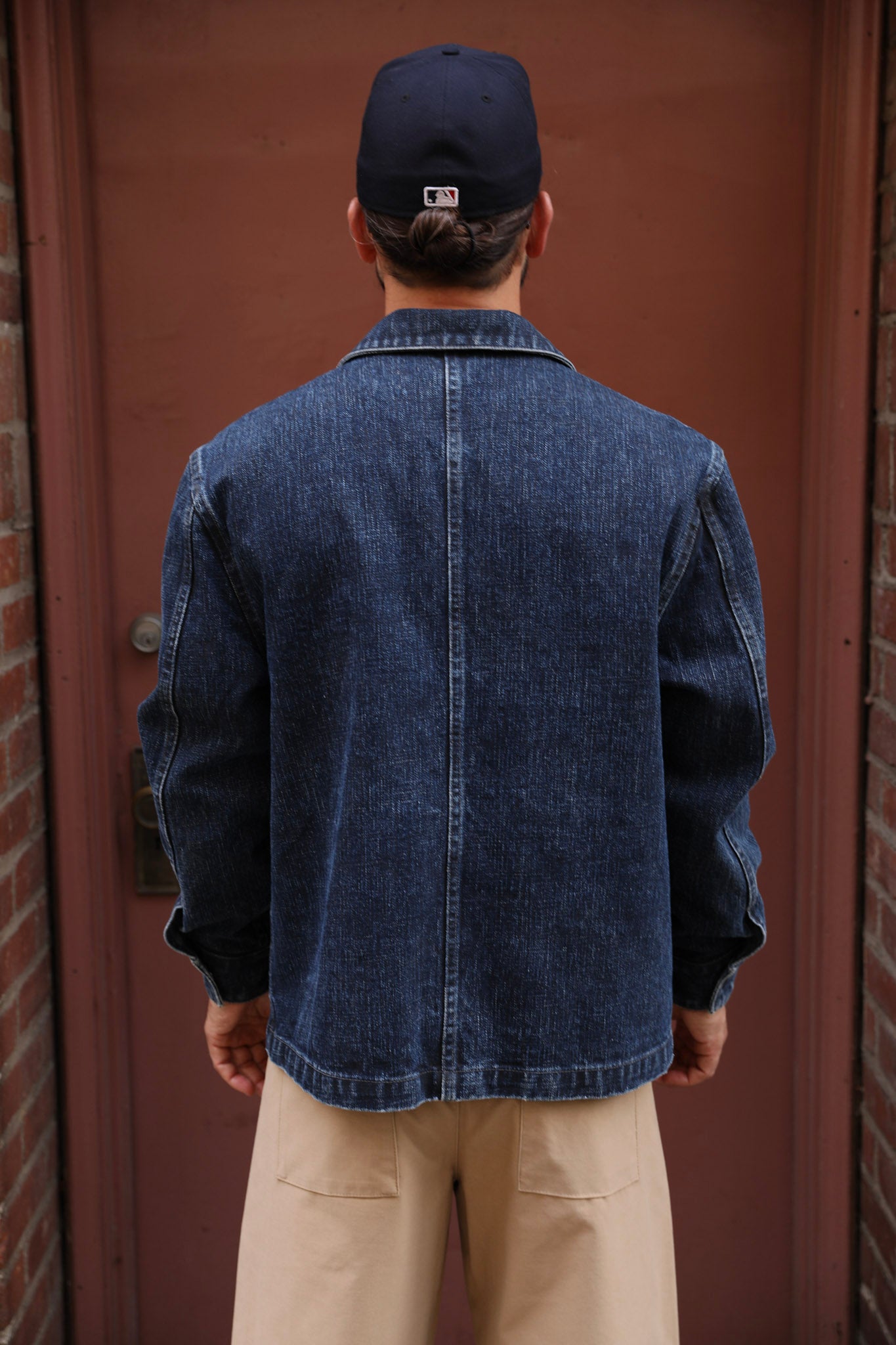 A man viewed from behind wearing a dark blue denim jacket, beige pants, and a navy baseball cap in front of a reddish-brown door.