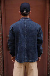 A man viewed from behind wearing a dark blue denim jacket, beige pants, and a navy baseball cap in front of a reddish-brown door.