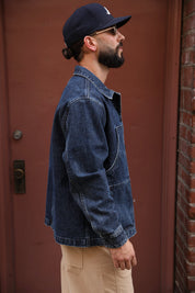 A man wearing a dark blue denim jacket, khaki pants, a navy baseball cap, and sunglasses, standing in profile against a reddish-brown door and brick wall.