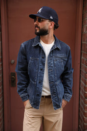 A man wearing a dark blue denim jacket over a white t-shirt, khaki pants, and a navy baseball cap with a white 'A' logo, standing in front of a reddish-brown door.