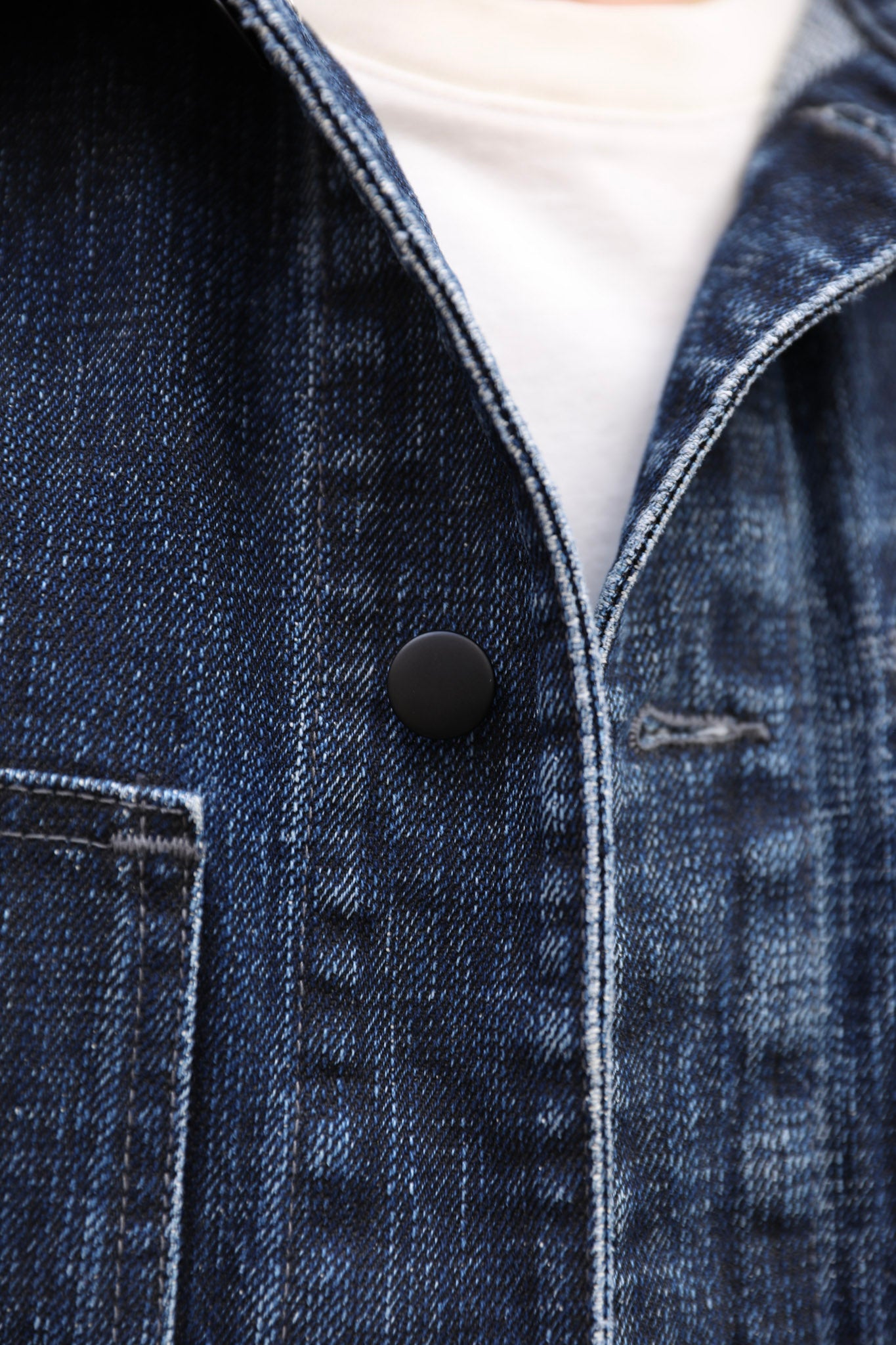Close-up of a dark indigo denim jacket with a black snap button, showing the textured denim weave and white contrast stitching over a white t-shirt.