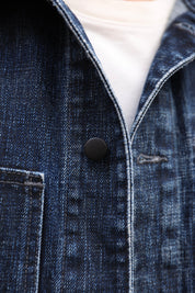 Close-up of a dark indigo denim jacket with a black snap button, showing the textured denim weave and white contrast stitching over a white t-shirt.