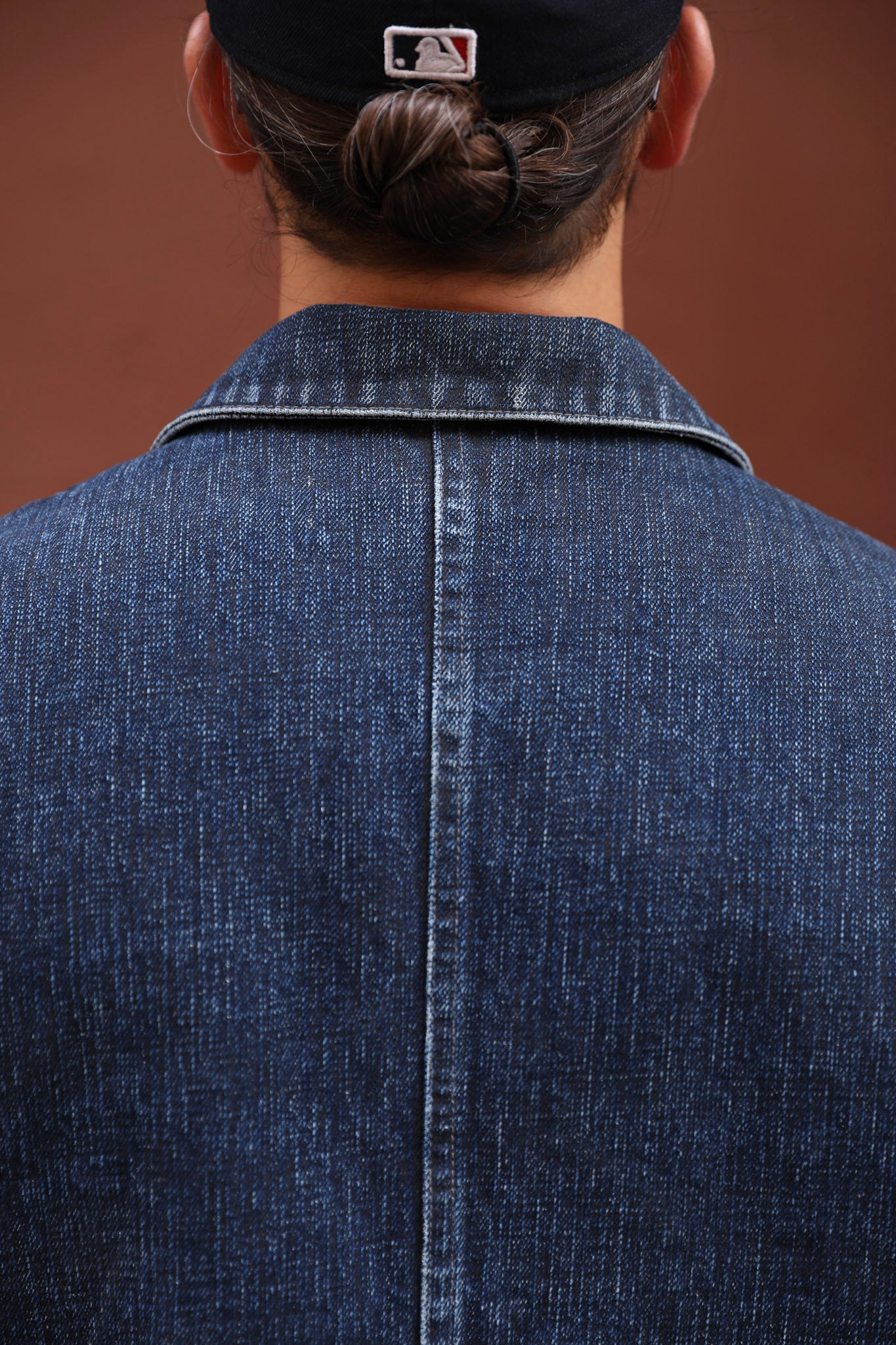 Close-up back view of a person wearing a dark indigo denim jacket and a black baseball cap with a visible MLB logo, against a warm brown background.