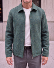 A man wearing a textured sage green zip-up jacket over a white t-shirt and gray trousers, standing outdoors against a light stone building.