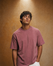A man wearing a washed, muted dusty rose short-sleeve t-shirt and white pants, standing against a textured warm brown studio backdrop.